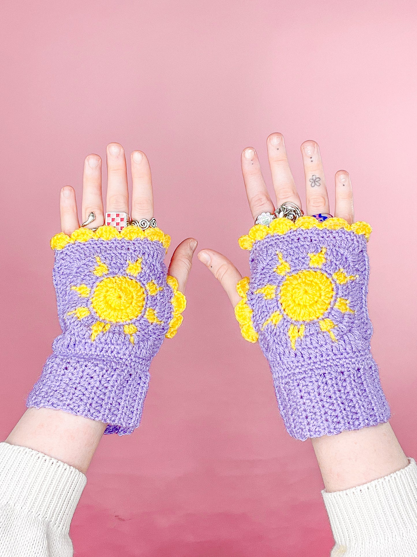 A pair of hands wearing granny square gloves in purple with a sun design and paired with various styles of rings.