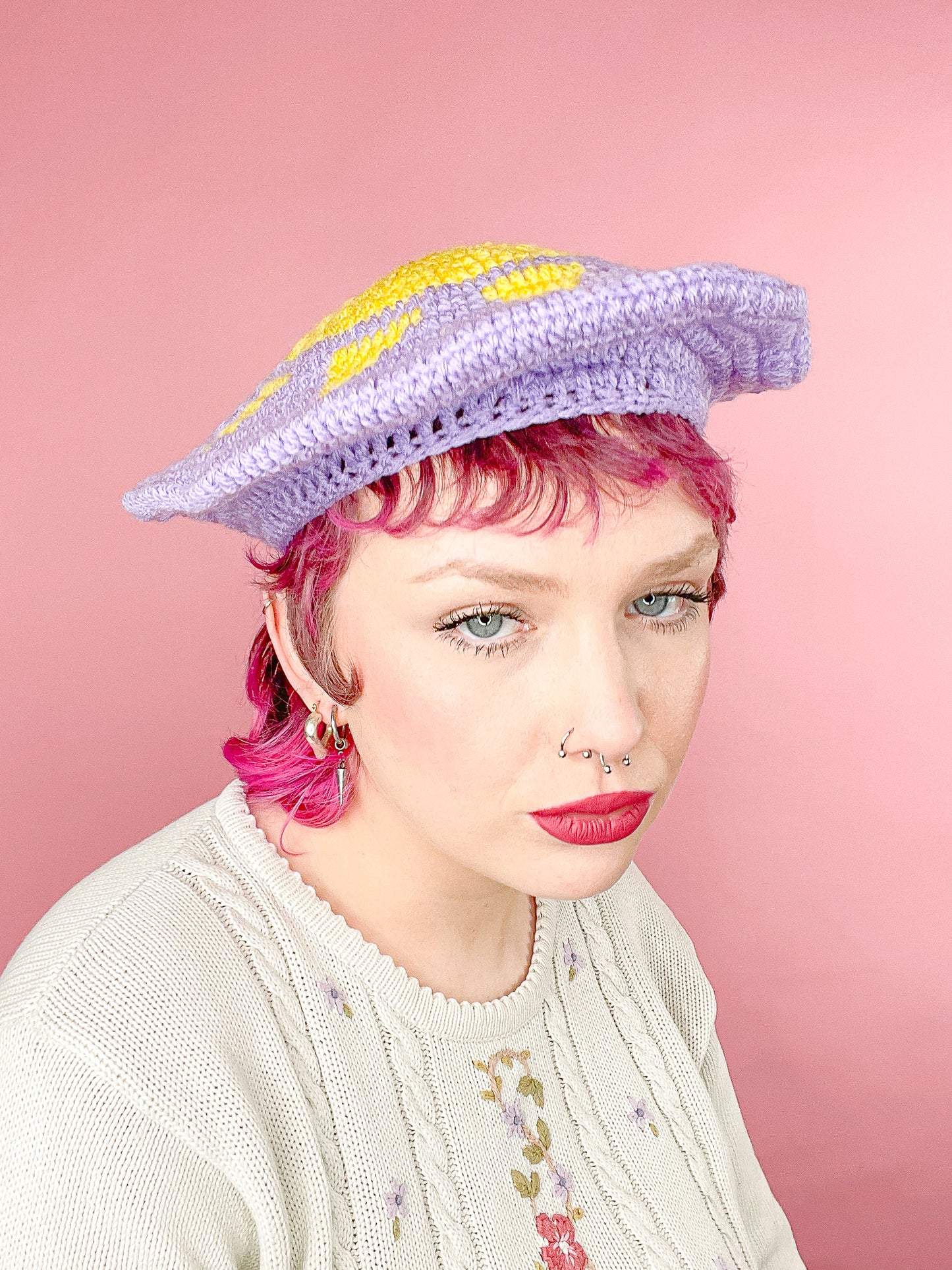 Front view of a woman wearing a lilac crochet hat with a yellow sun on the top paired with a white granny style jumper.