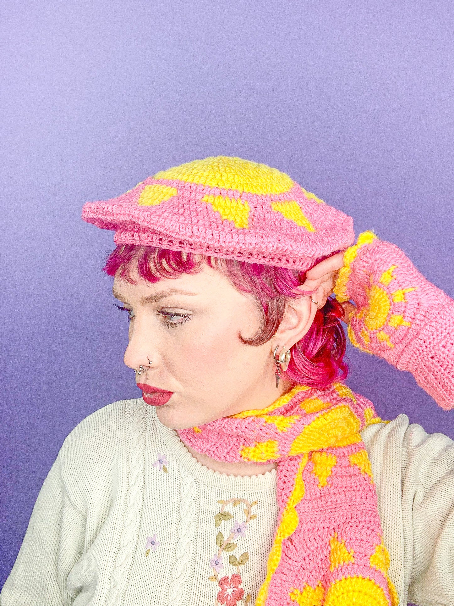 Side view of a woman wearing a pink beret with a sun on the top paired with a matching crochet scarf and fingerless gloves.