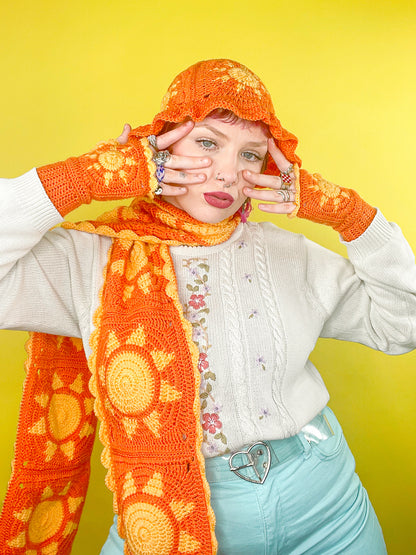 Front view of a woman posing wearing a crochet set of an orange scarf, gloves and bucket hat with a yellow sun design and contrasting edges.