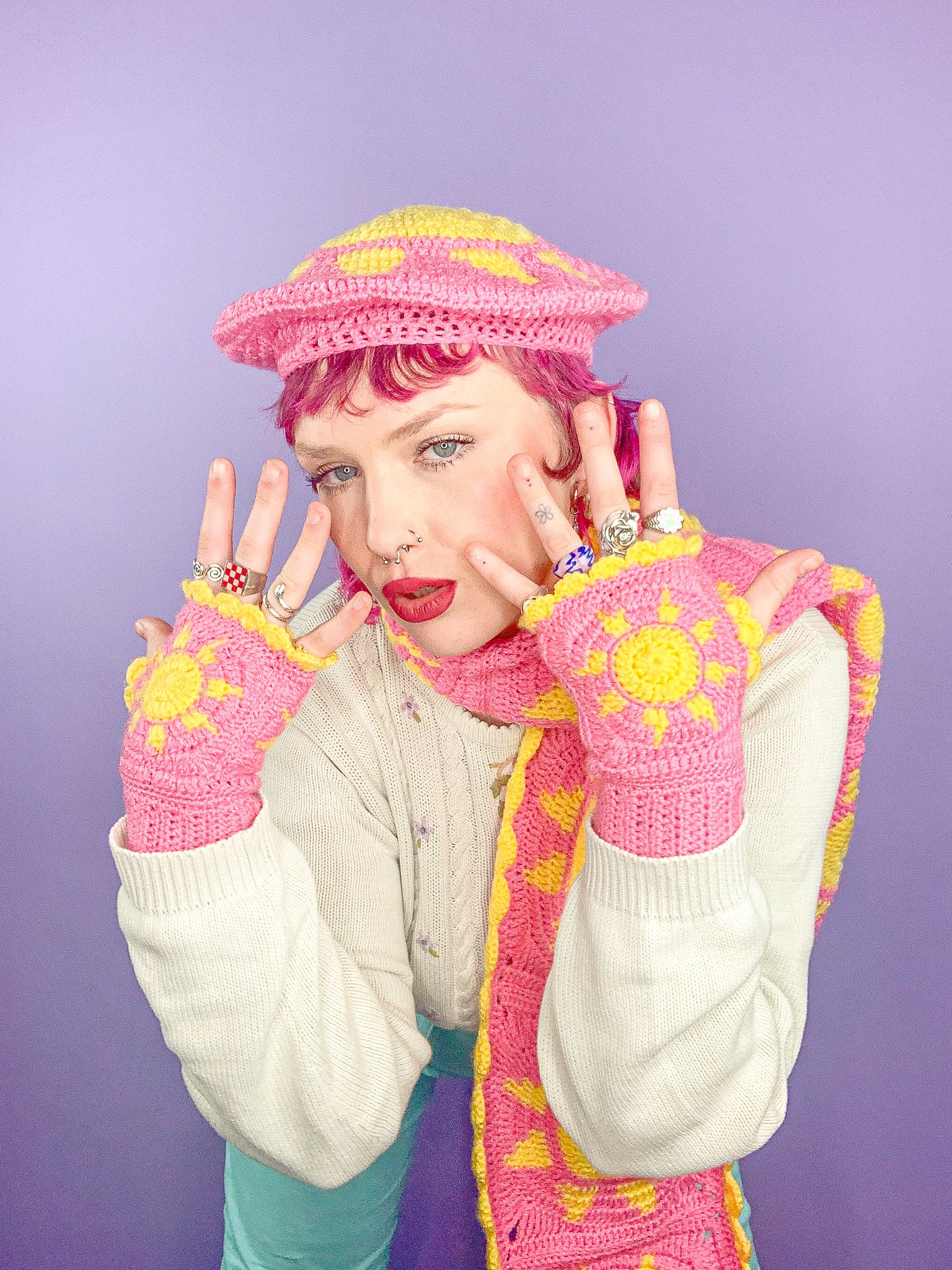 Close view of woman posing with her hands in front of her face wearing a matching set of pink gloves, a beret hat and scarf with suns all over them.