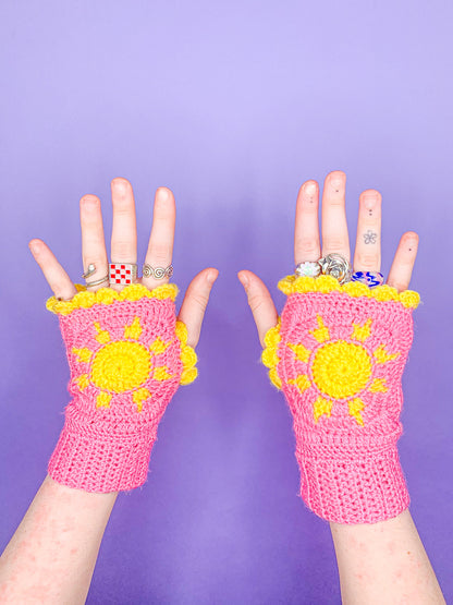 A pair of hands facing upwards wearing granny square gloves in pink with a yellow sun design and frilled edges.