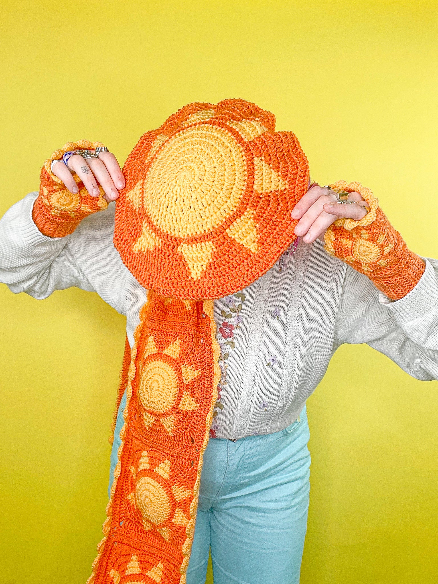 Above view of a woman wearing an orange beret with a sun on the base, paired with a matching granny square scarf and fingerless gloves.