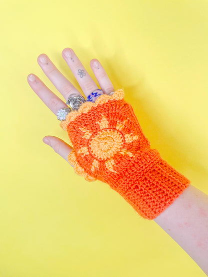 One hand wearing rings and an orange crochet glove with a sun on it and contrasting edges against a yellow background.