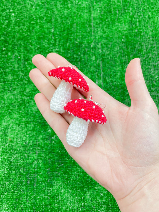 A hand holding a pair of crochet earrings shaped like toadstool mushrooms in red in front of a patch of grass.