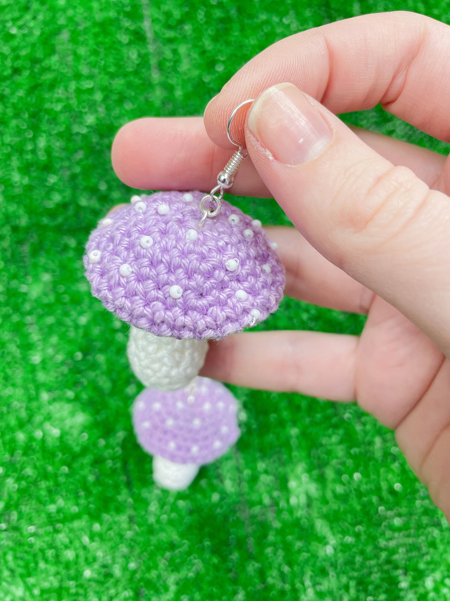 A hand holding on crochet earring shaped like a toadstool mushroom in lilac showing the white seed beads attached resembling mushroom dots.