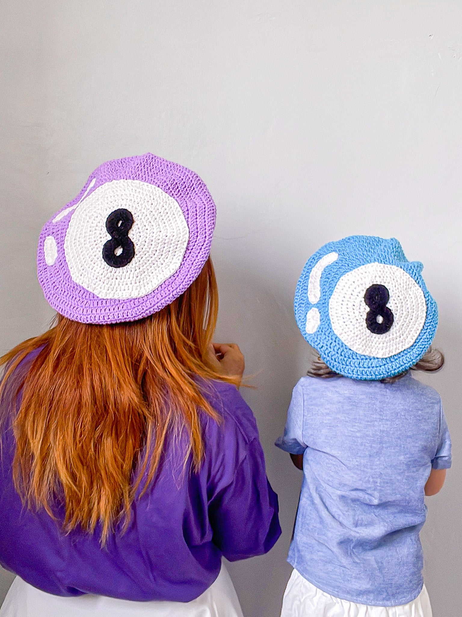 Back view of a mother and child wearing matching crocheted berets with 8 balls on them, the mum wearing lilac and the boy wearing baby blue.