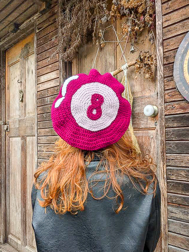 Back view of a woman wearing a beret hat in bright pink with an 8 ball design on it standing in front of a wooden cabin.