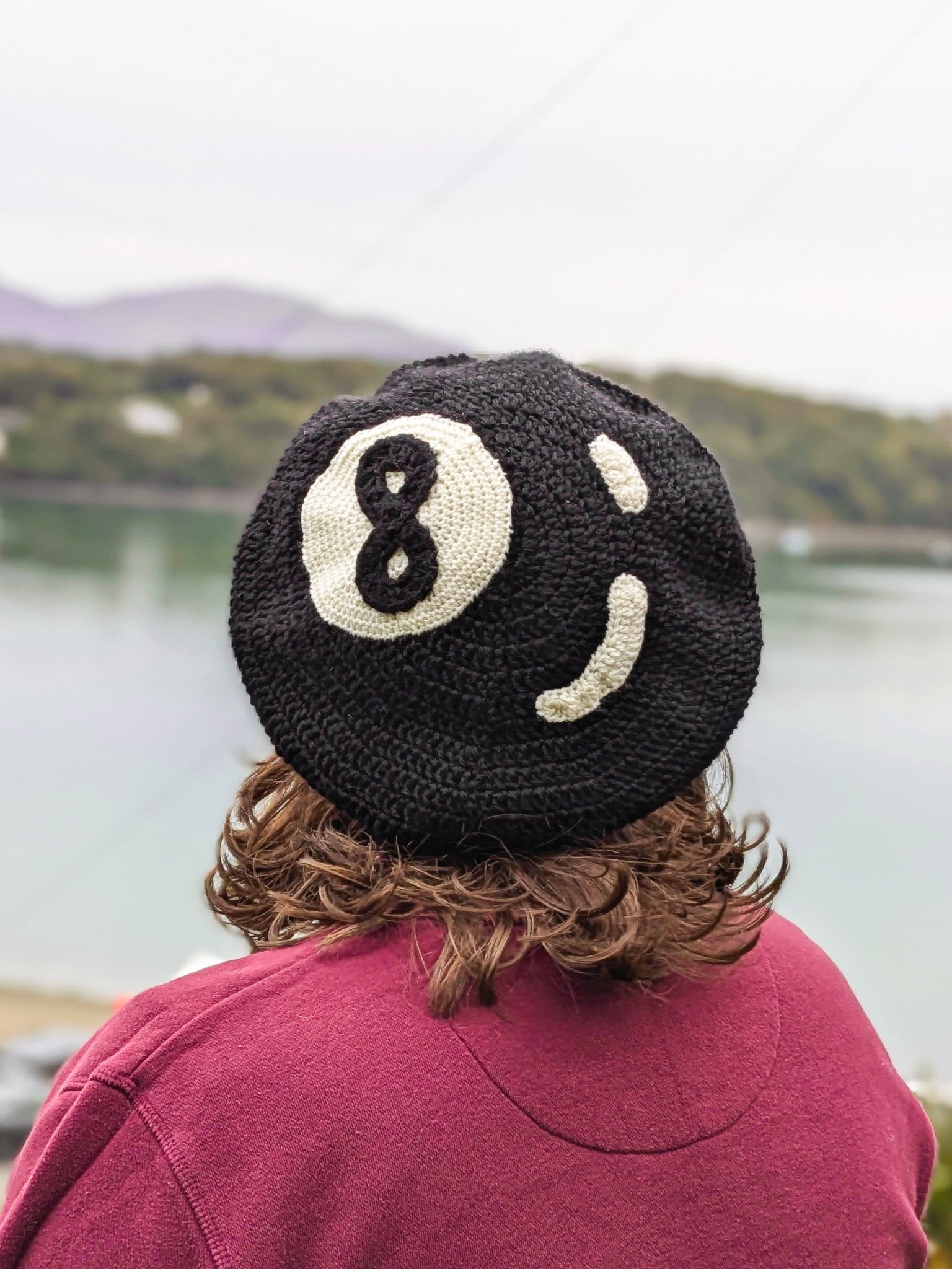 Back view of a person wearing a crocheted beret hat that resembles an 8 ball, standing in front of a river.