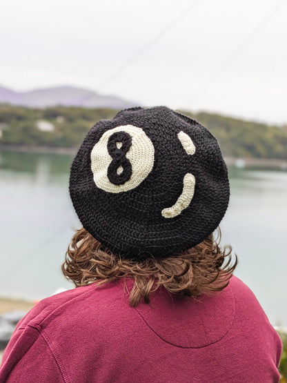 Back view of a person wearing a crocheted beret hat that resembles an 8 ball, standing in front of a river.