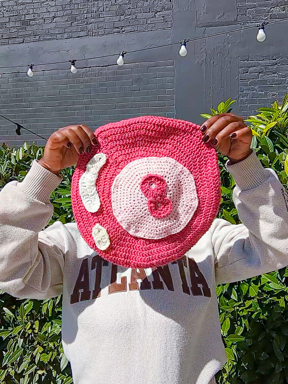 A woman holding a pink crochet beret that resembles an 8 ball in front of her face.