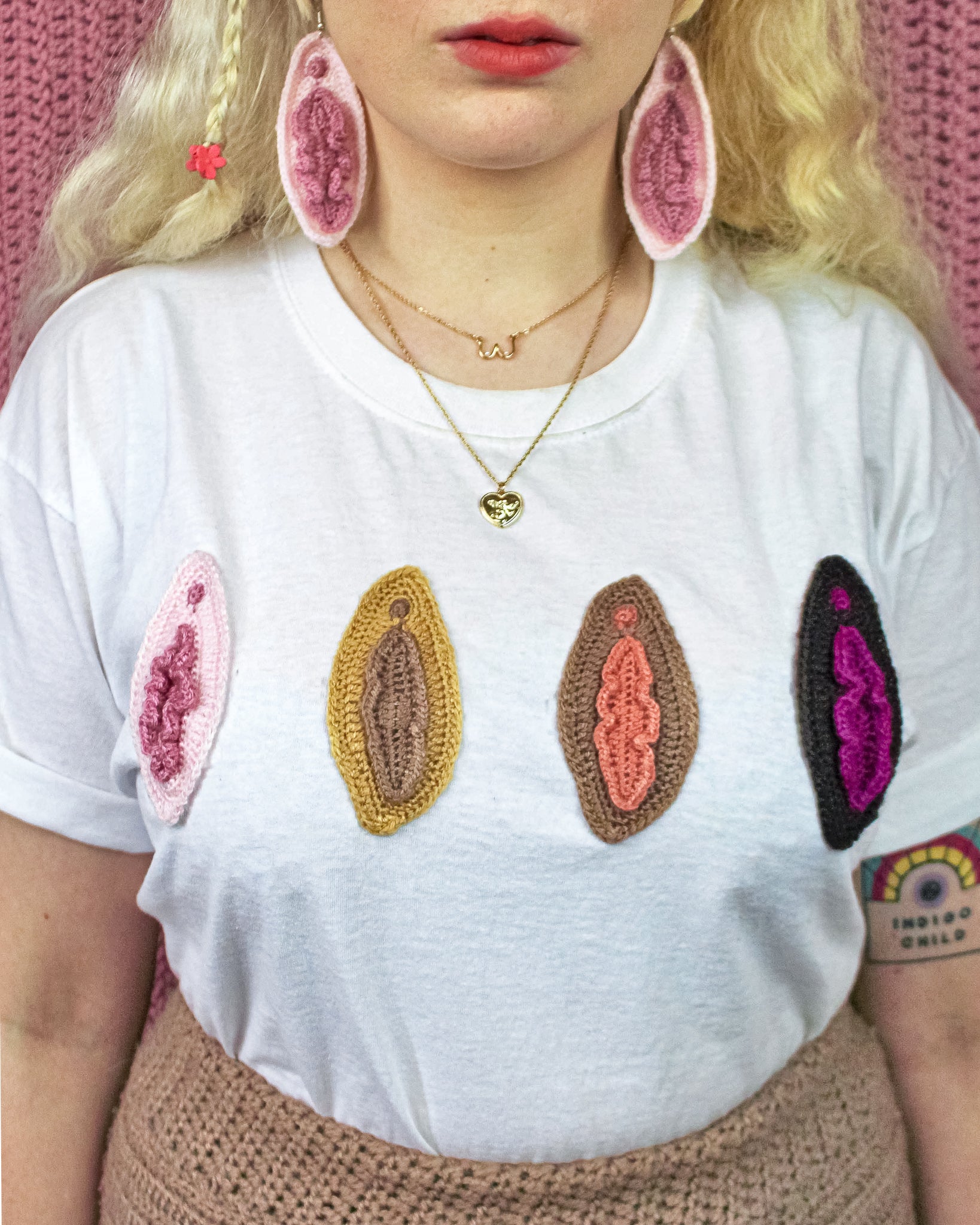 Close up of a woman wearing a white t-shirt with four crocheted vulvas sewn onto the front in a row. Each vulva is a different skin tone from cream, tan, light brown and dark brown with matching vulva earrings.