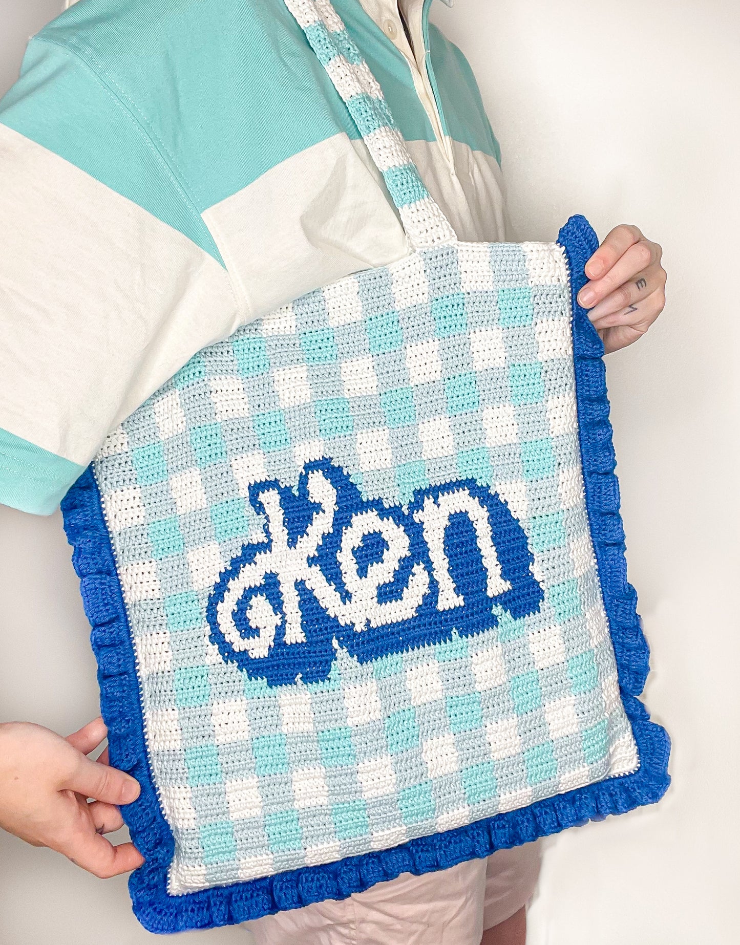 Close view of a person wearing a crochet shoulder bag with blue and white gingham and the word 'Ken' on the front, paired with a matching blue and white stripe polo.