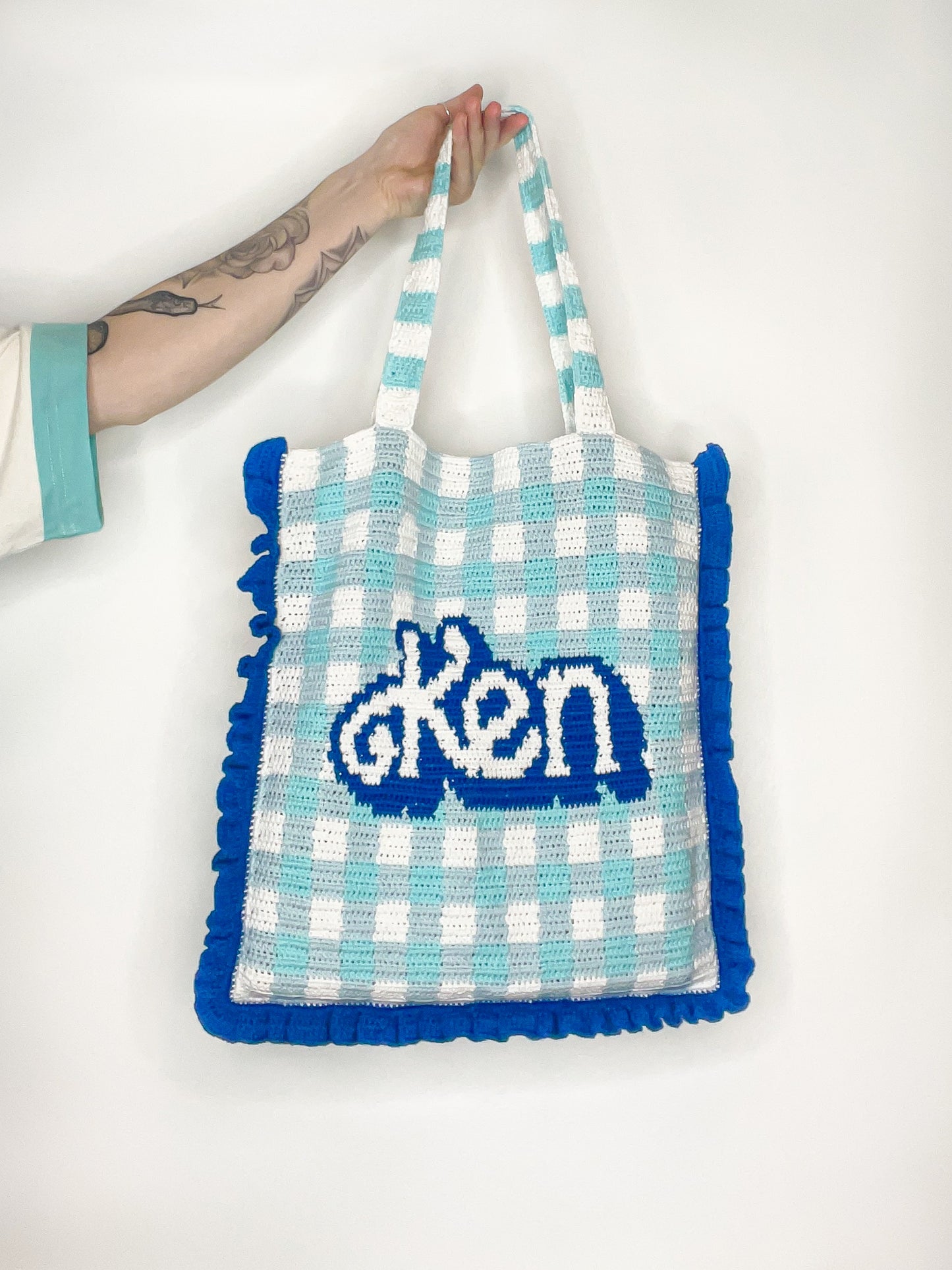 A hand holding a blue and white tote bag with dark blue frills and 'Ken' on the front, held in front of a white wall.