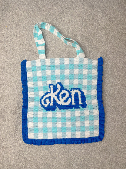 Flat lay of a blue and white gingham tote bag with 'Ken' on the front and a dark blue frilly edge lying on a grey speckled rug.