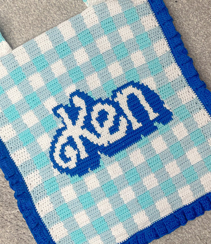 Close view of a crochet tote bag in baby blue and white gingham with dark blue frills and 'Ken' on the front showing the crochet stitches.