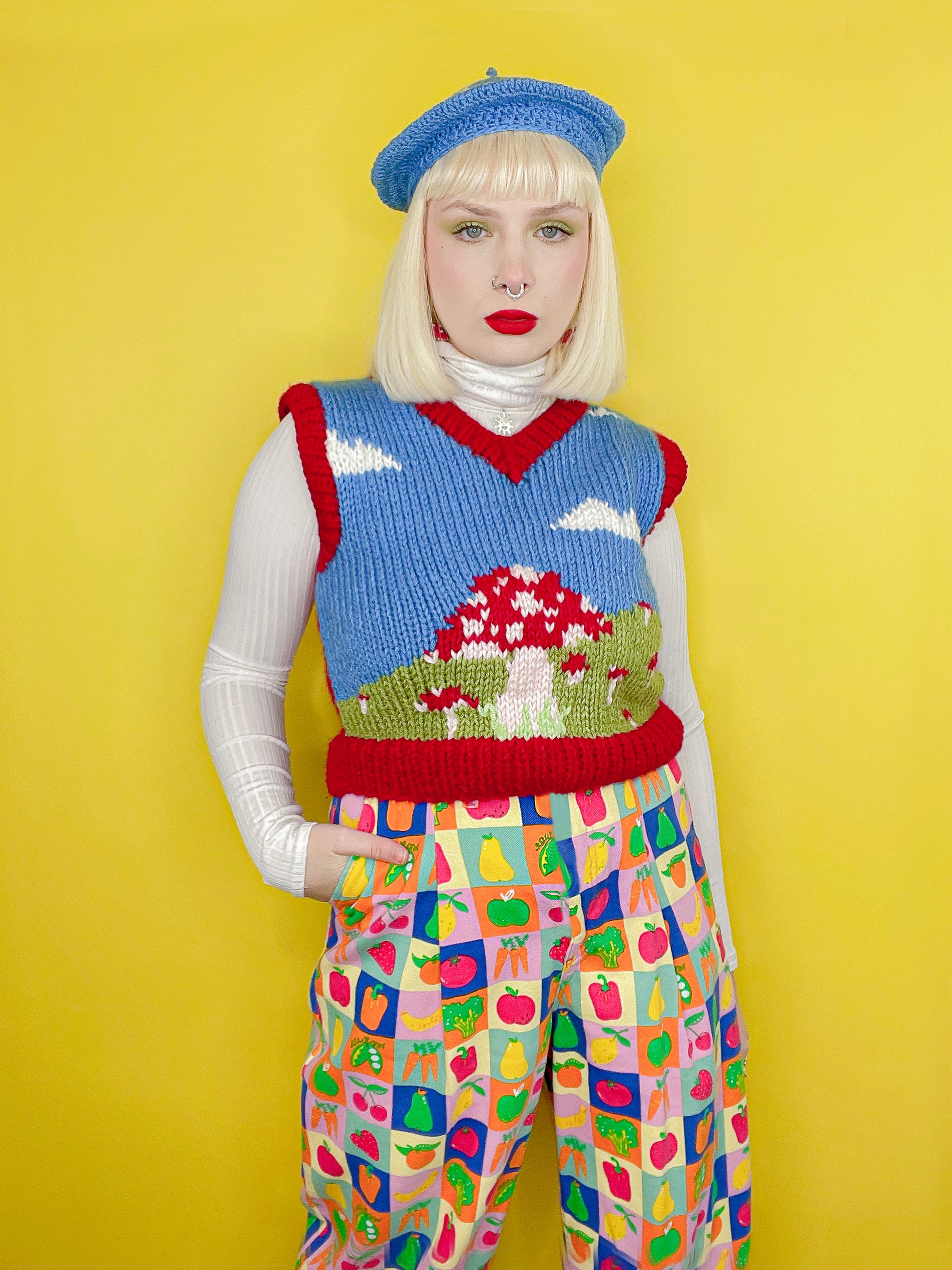 A woman posing wearing a mushroom motif vest top with a dark blue cloud background and red edges, paired with fruit and vegetable jeans, a white turtleneck and blue beret.