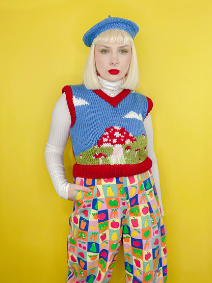 A woman posing wearing a mushroom motif vest top with a dark blue cloud background and red edges, paired with fruit and vegetable jeans, a white turtleneck and blue beret.