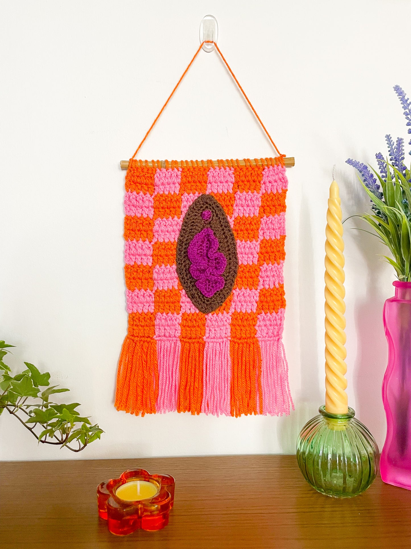 A wall hanging in orange and pink with dark brown crochet artwork resembling a vulva on the front hung on a white wall with decor around it.