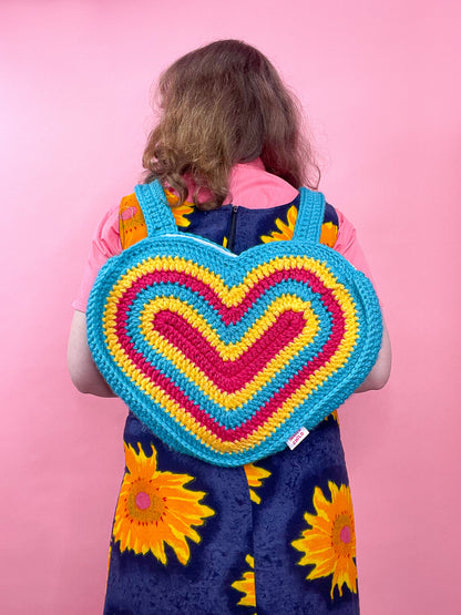 Back view of a woman wearing a crochet bag shaped like a heart in pansexual flag colours (pink, yellow, aqua blue), paired with a pink shirt and blue and yellow dress.