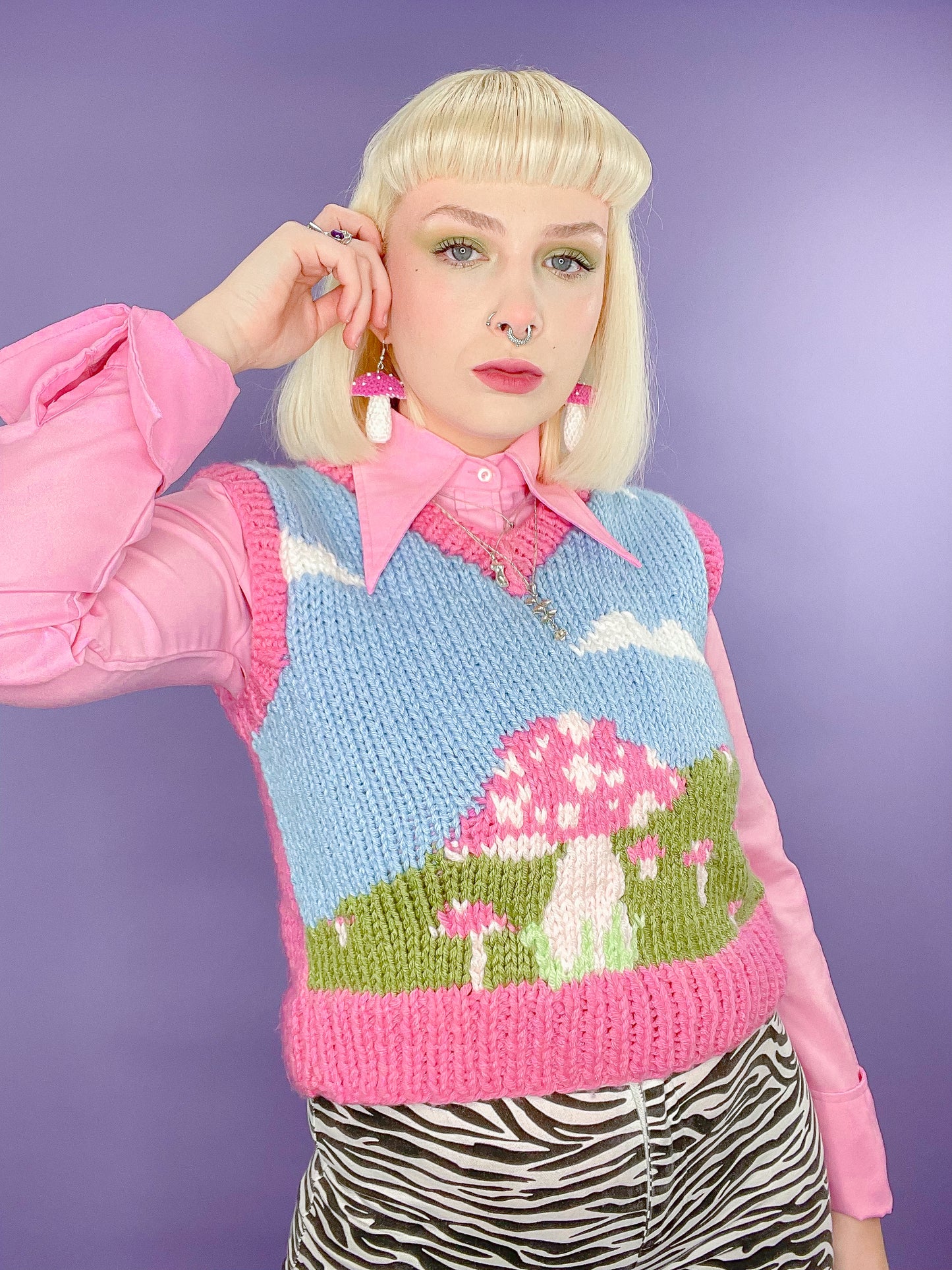 A woman posing wearing a knitted vest top with a mushroom field and clouds design with pink edges and a pink shirt underneath standing in front of a purple background.