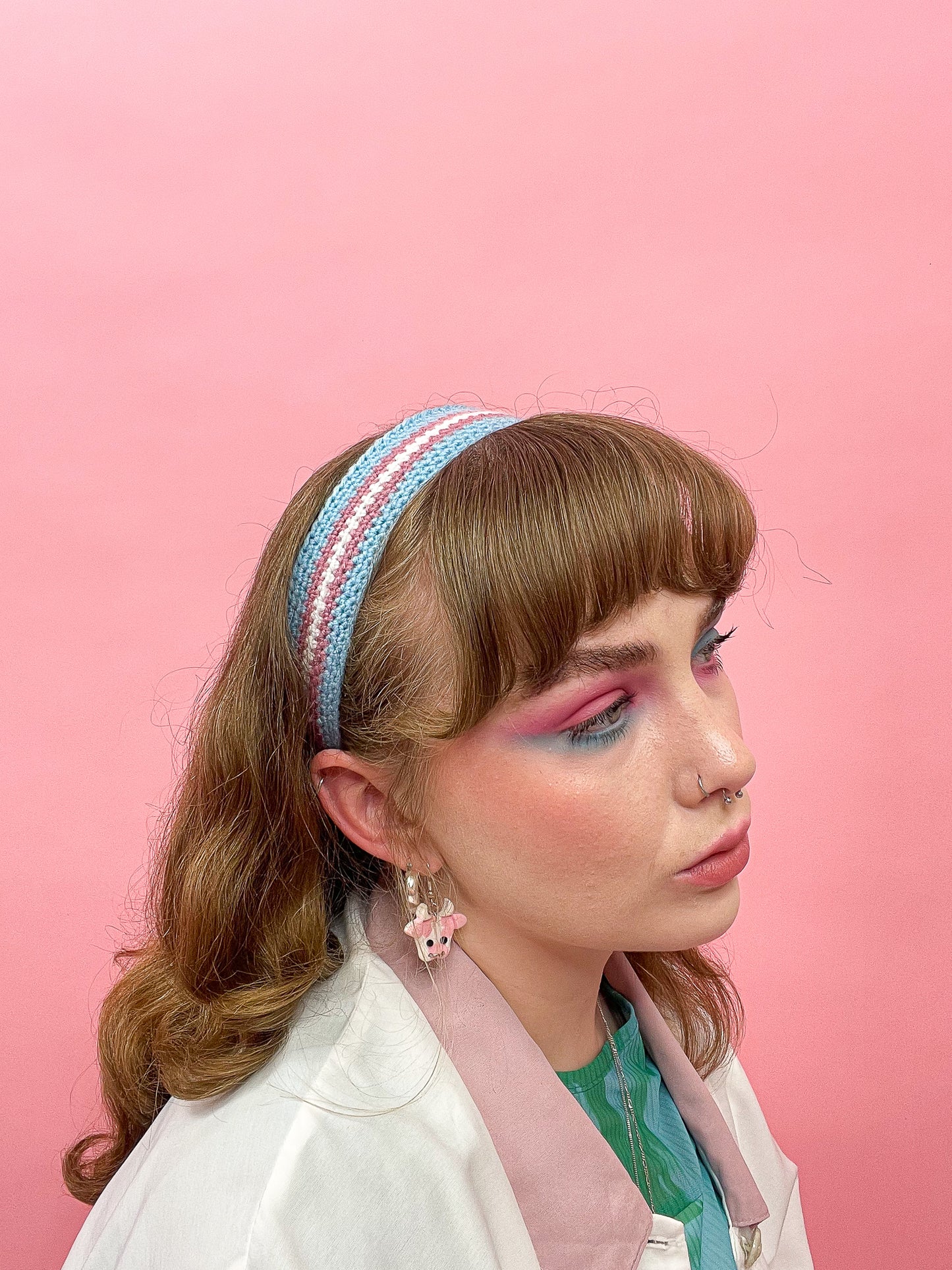 Side view of a woman wearing a crochet headband resembling the trans flag colours (blue, pink, white), with a matching outfit and eyeshadow.