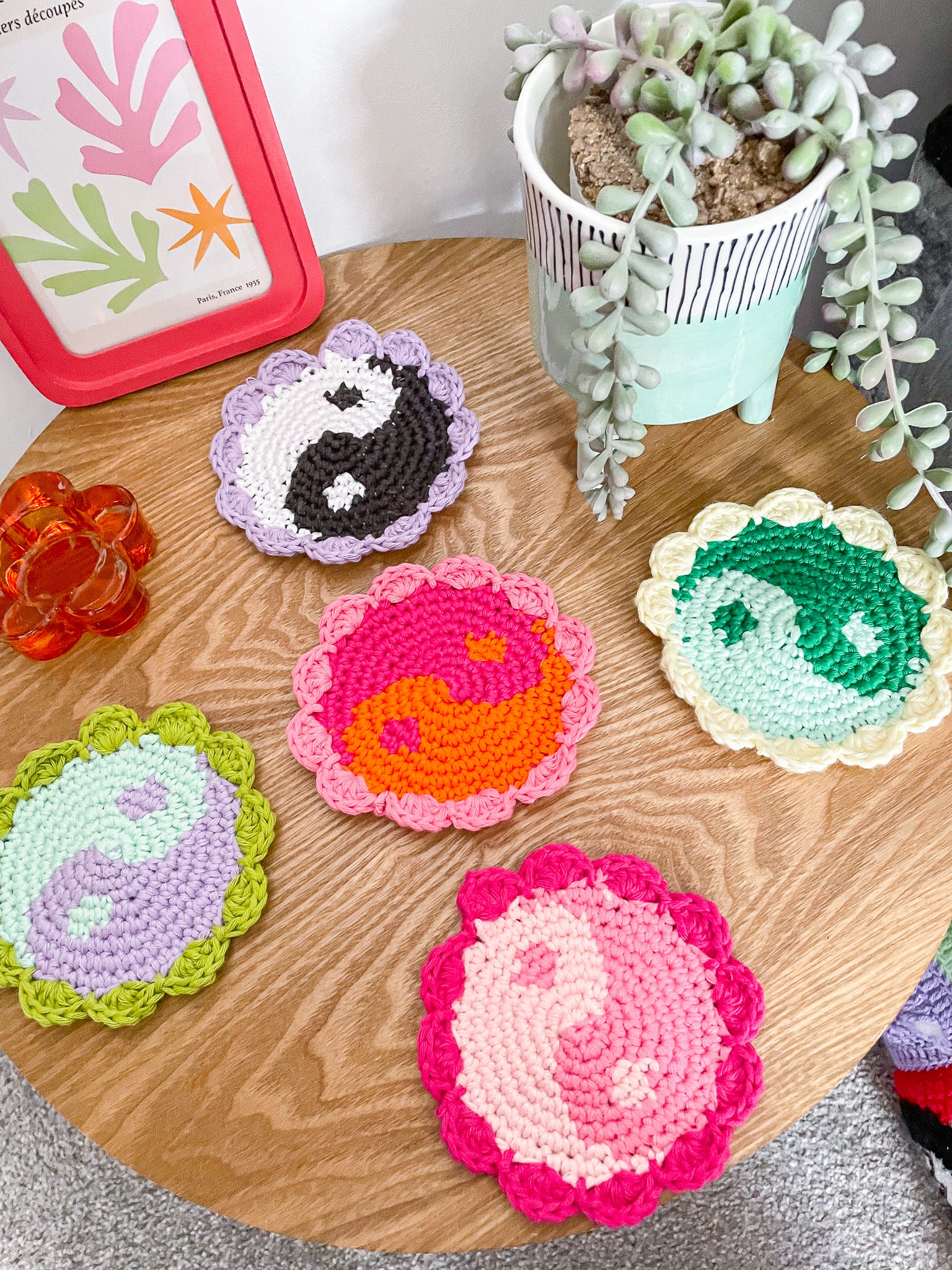 Five crochet yin yang coasters in various different colours sat together on a wooden coffee table in an X shape with home decor around them.