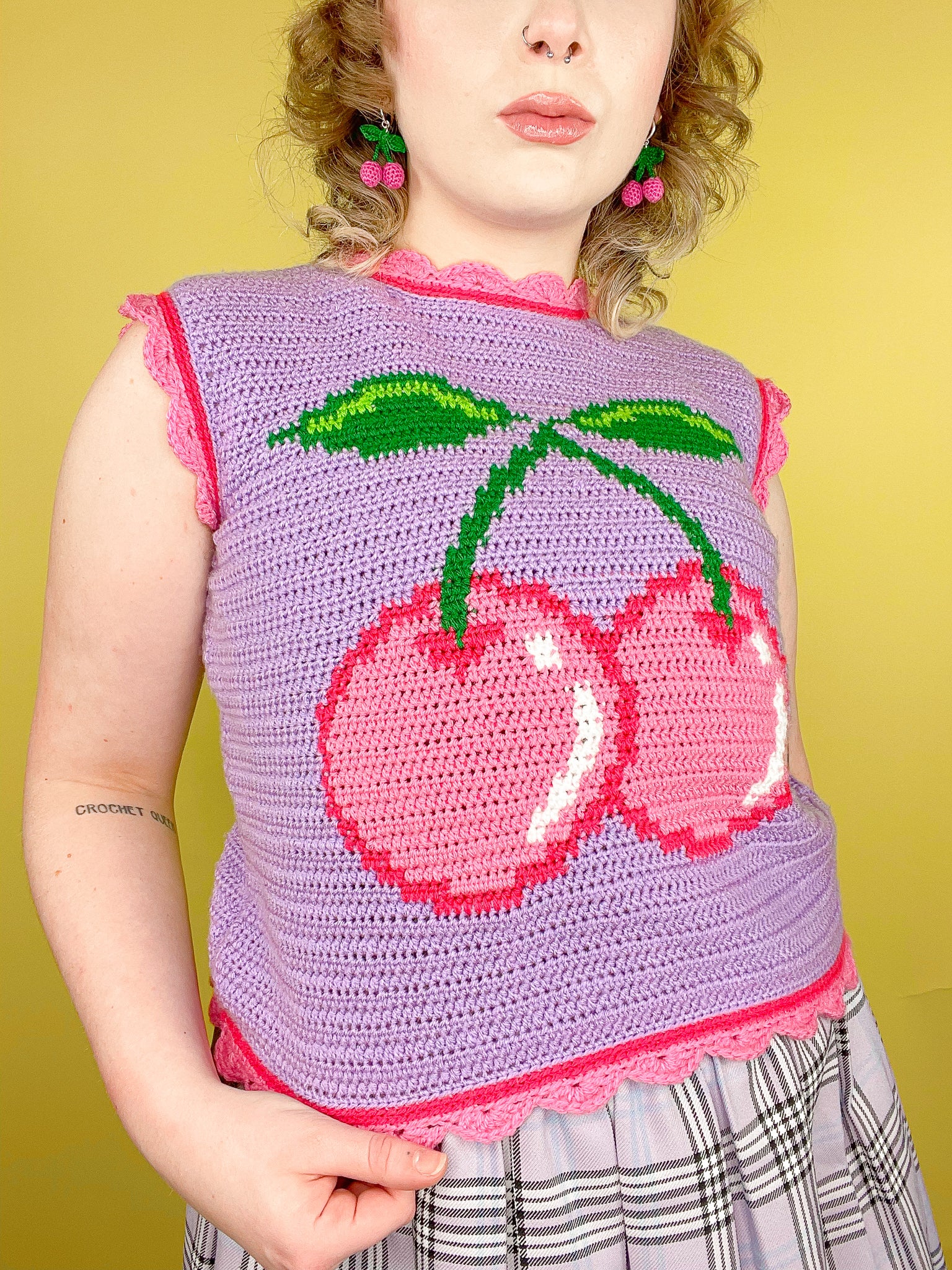 Close view of woman wearing a crochet vest in lilac with a pink cherry on the front and frills with a matching purple kilt.