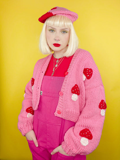 A woman posing wearing a hand knit crop cardigan in pink with an appliqué red mushroom design, paired with a pink and red beret, a red tank top, pink overalls and red lipstick.