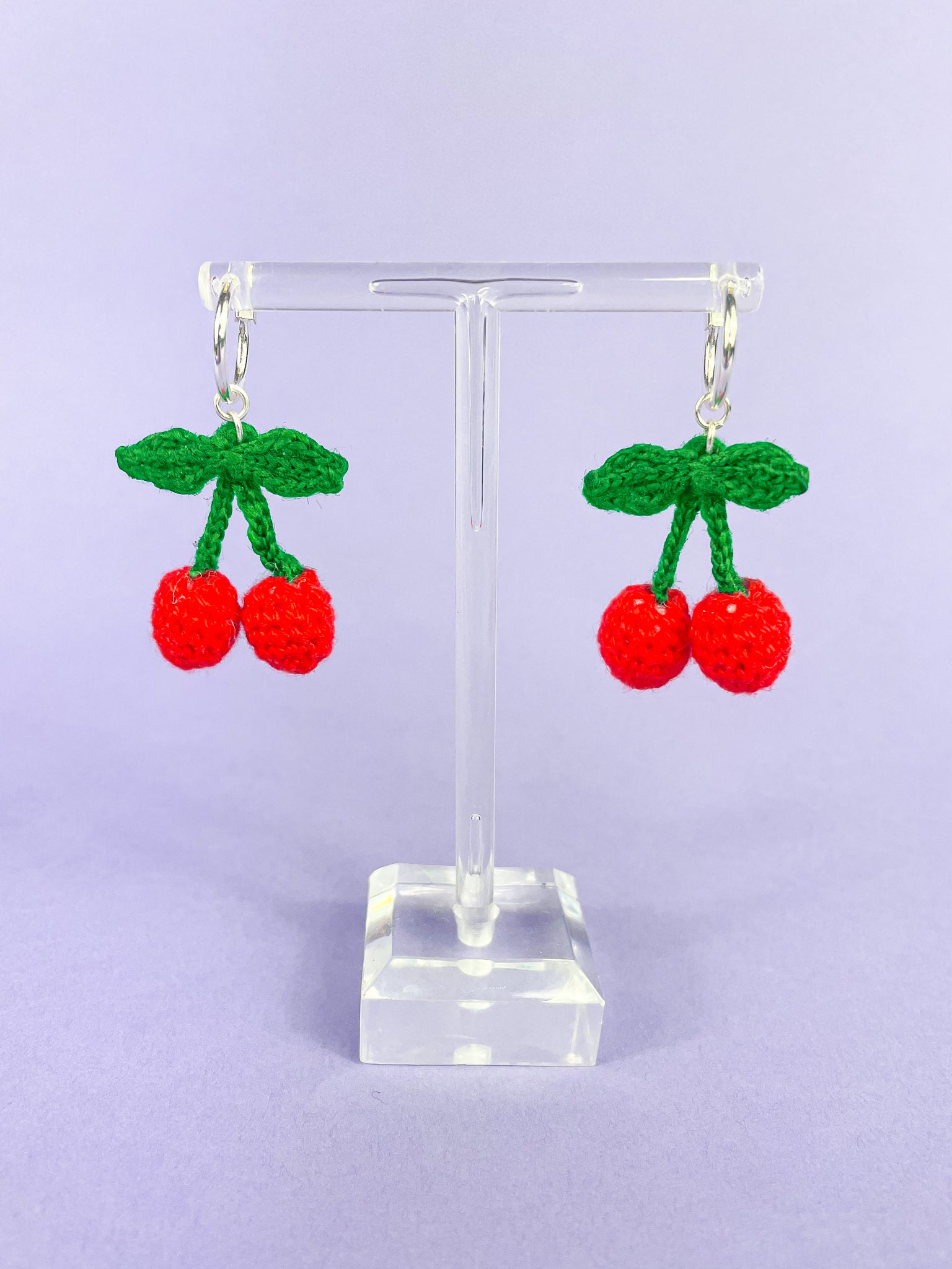 A pair of silver hoop earrings with crocheted red cherries attached to them hanging on an earring stand against a purple background.