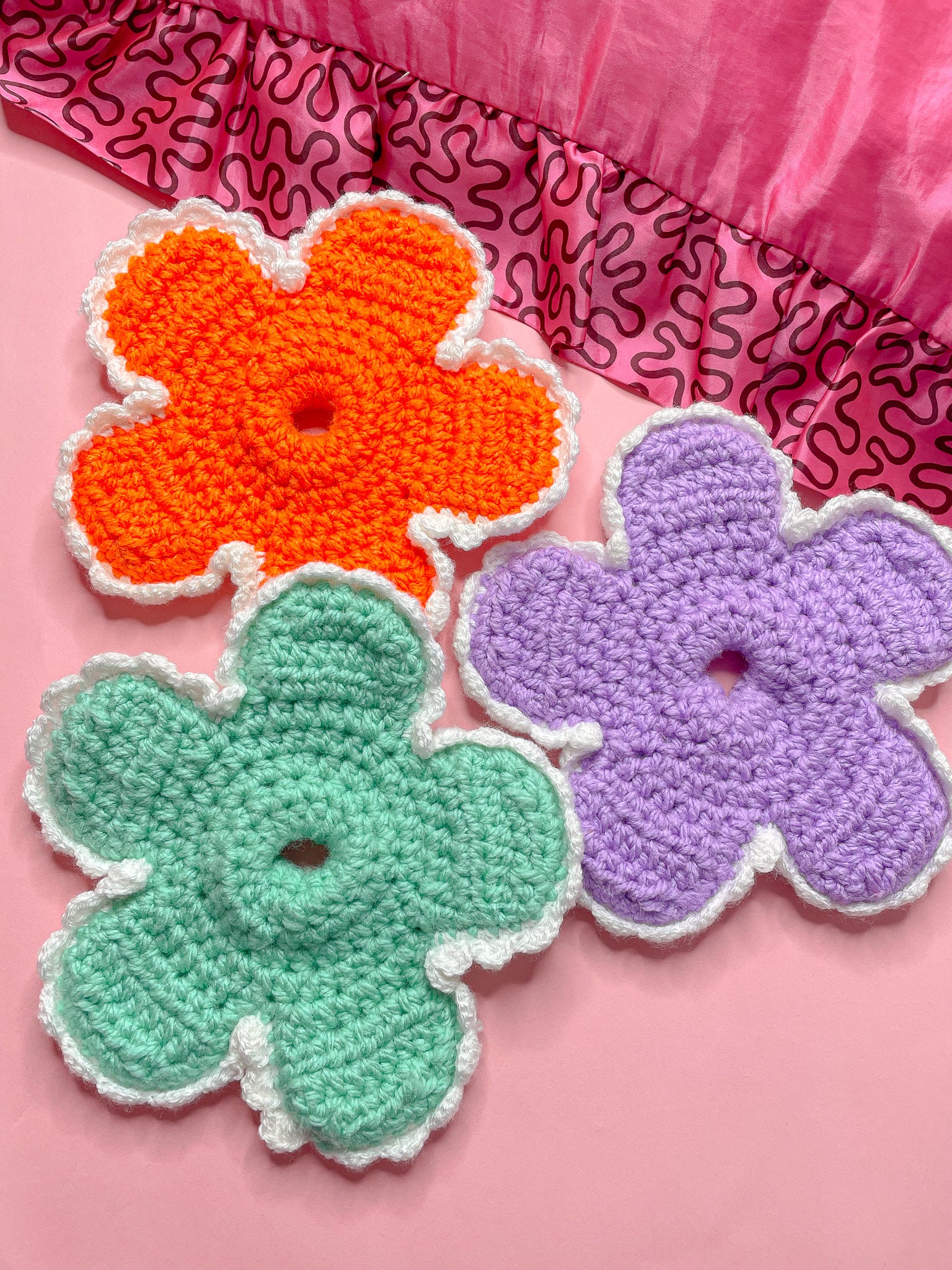 Three crochet scrunchies shaped like daisies in orange, lilac and mint green lying together on a pink background.