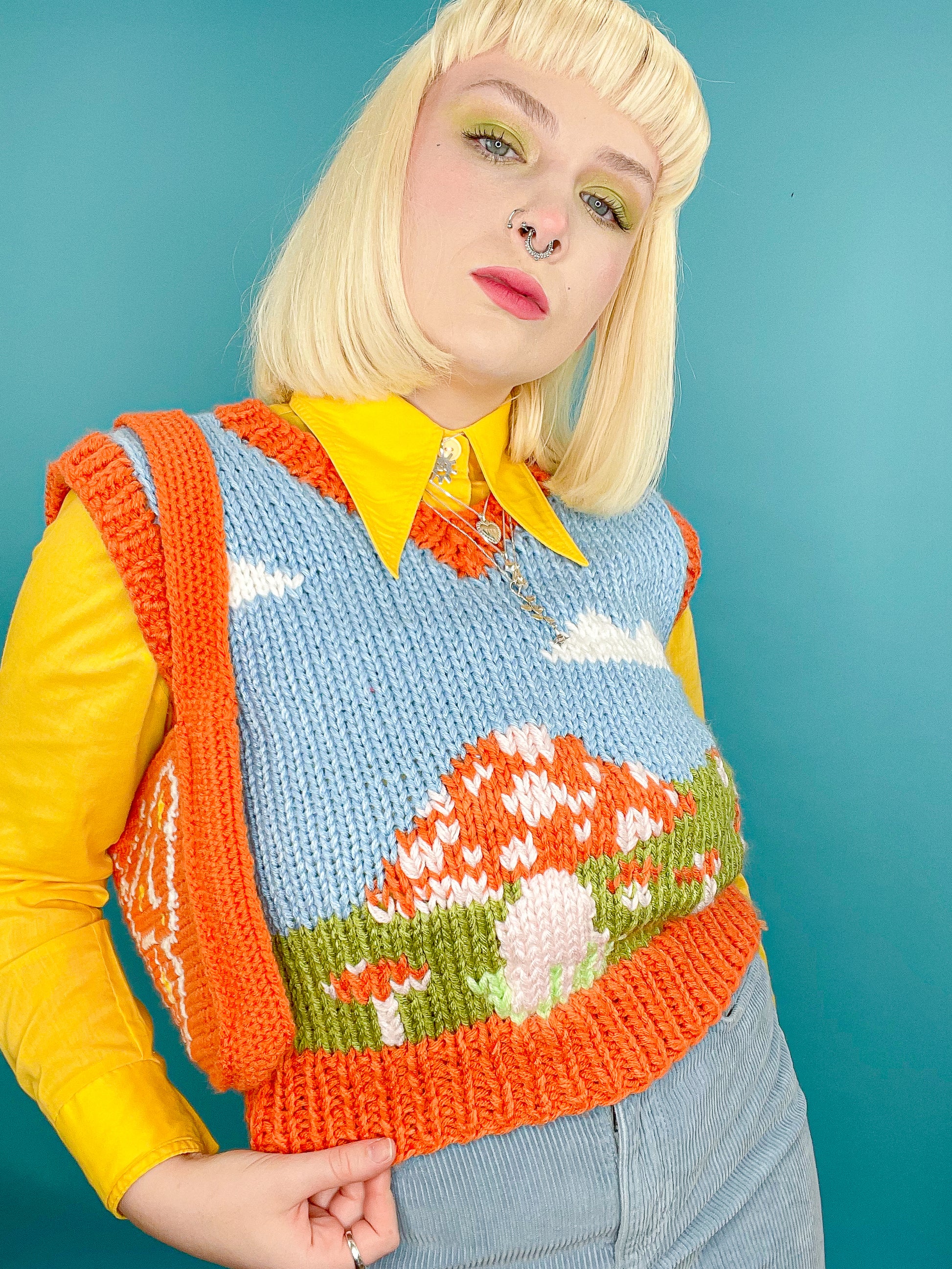 Close up of woman posing wearing a baby blue and orange knitted vest with a field design and a matching shoulder bag with a yellow 70's shirt underneath.