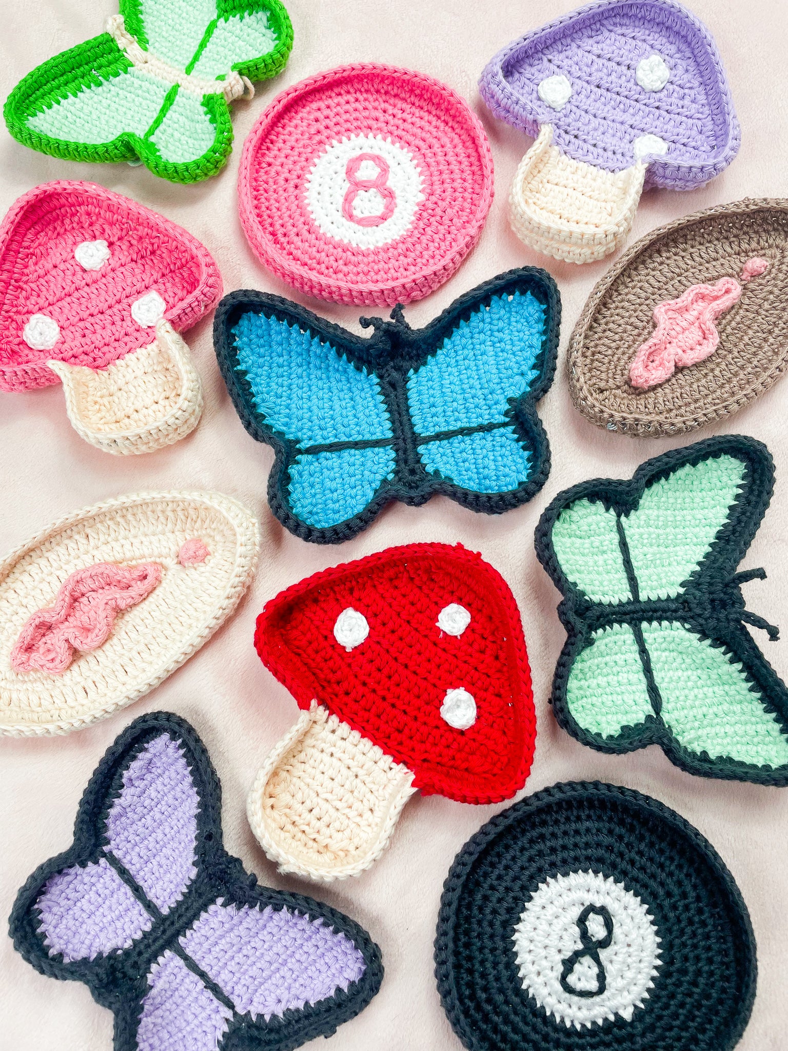 Eleven crochet trinket dishes all in different shapes (butterflies, mushrooms, 8 balls and vulvas) lying together on a pink background.