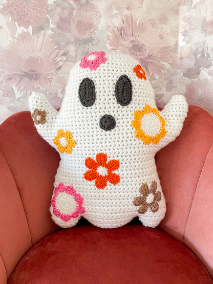 A ghost shaped pillow with pink, orange and brown flowers attached to it sitting on a pink velvet chair with a floral wallpaper in the background.