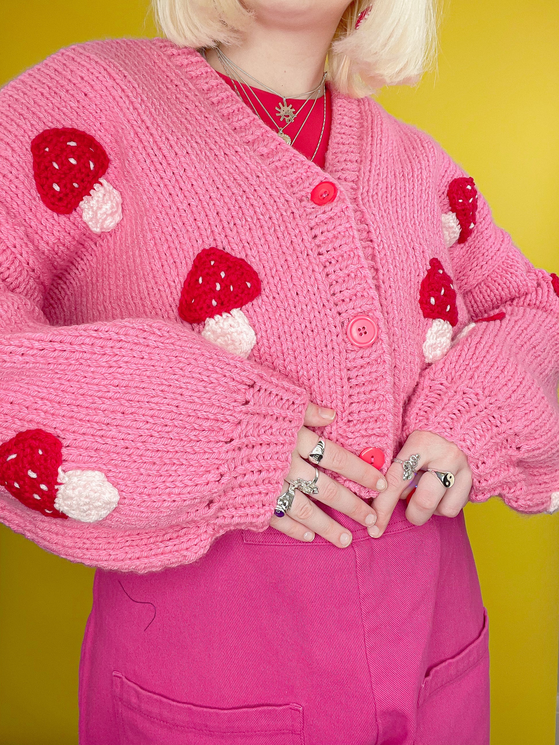 Close up of woman posing wearing a knitted cardigan in pink with red mushrooms all over and multiple rings.