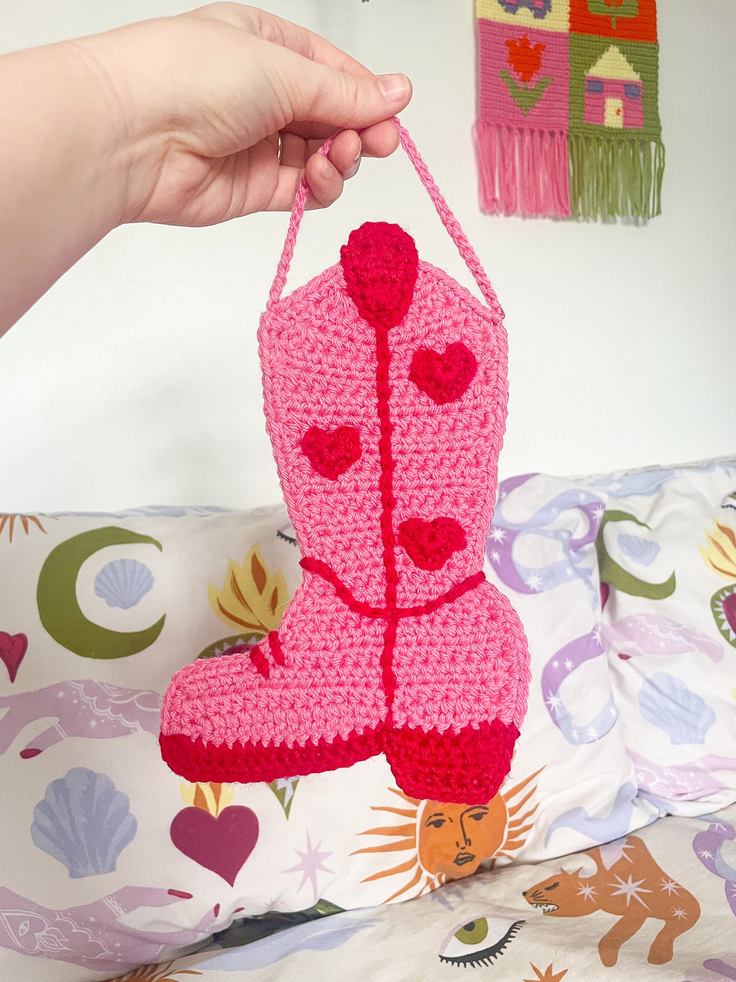 A hand holding a crochet wall hanging shaped like a cowboy boot in pink with red details in front of a patterned bed sheet and white wall.