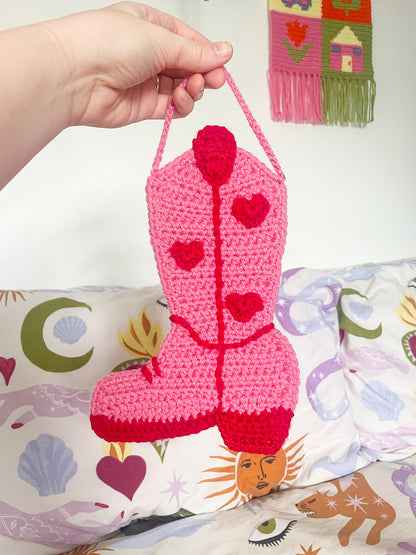 A hand holding a crochet wall hanging shaped like a cowboy boot in pink with red details in front of a patterned bed sheet and white wall.