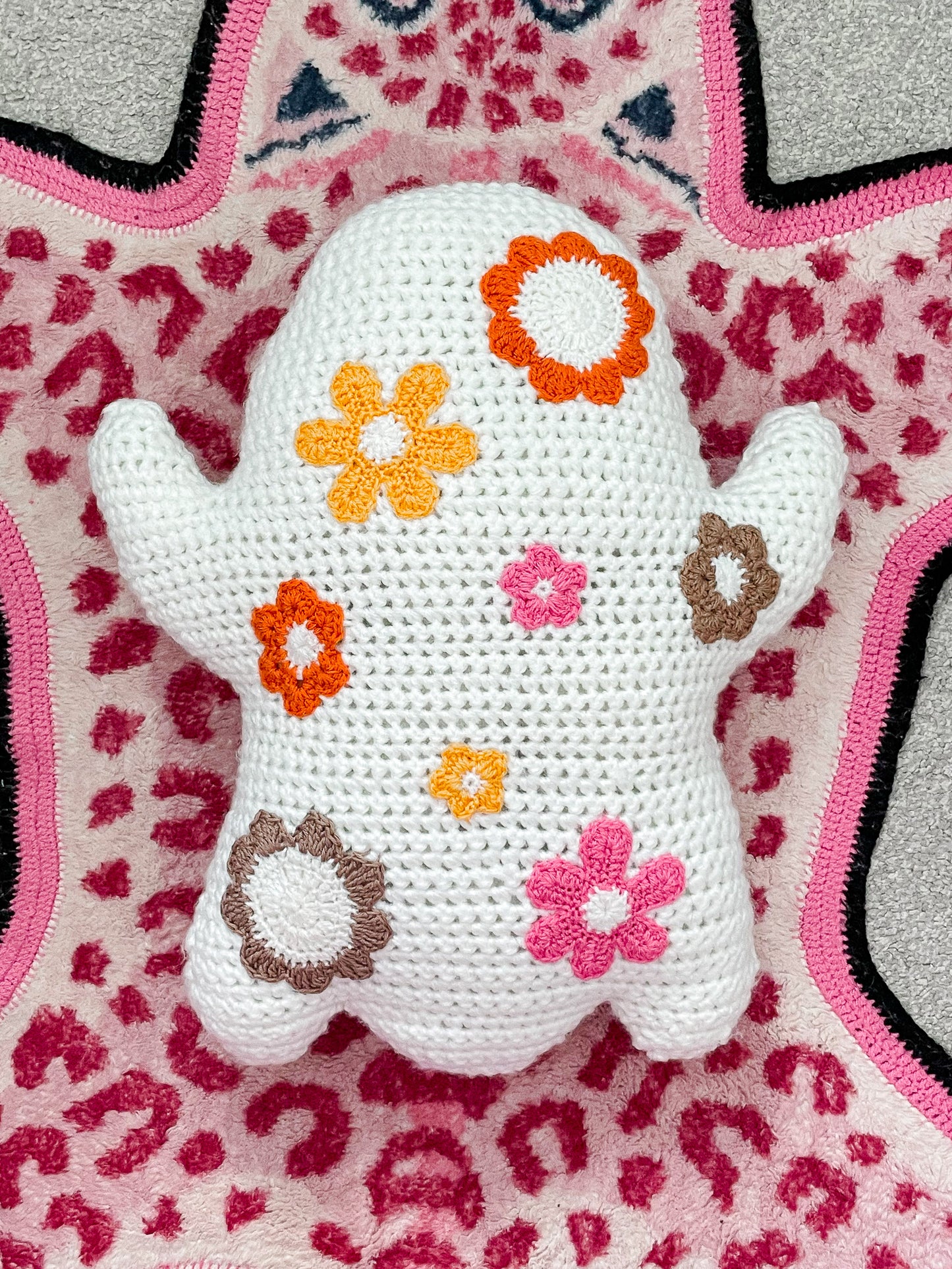 Back view of a white crochet pillow shaped like a ghost with multicoloured flowers sewn on it, lying flat on a pink leopard print rug.