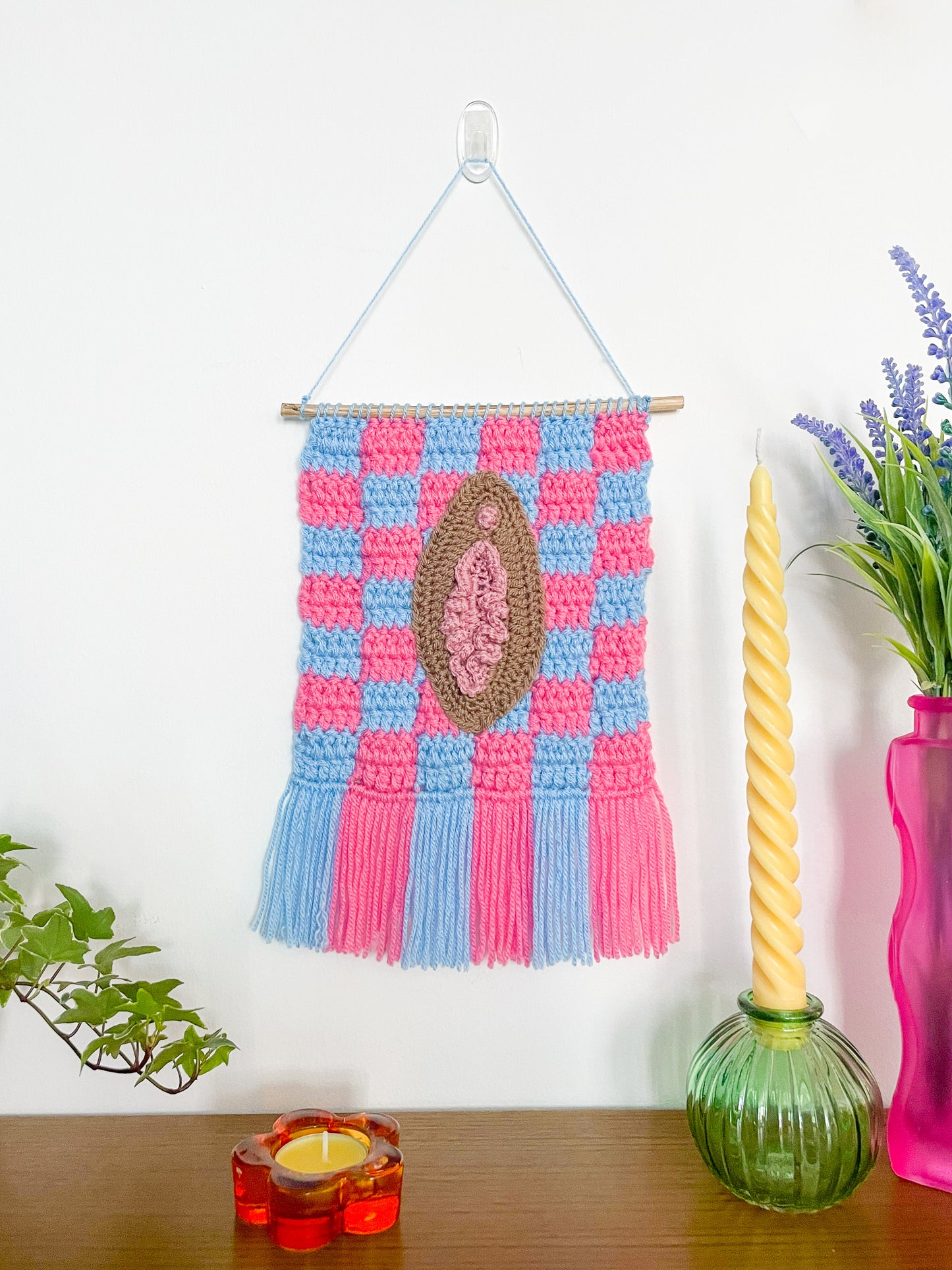 A blue and pink crochet wall hanging with a brown vulva sewn on the front hung on a white wall with candles and plants around.