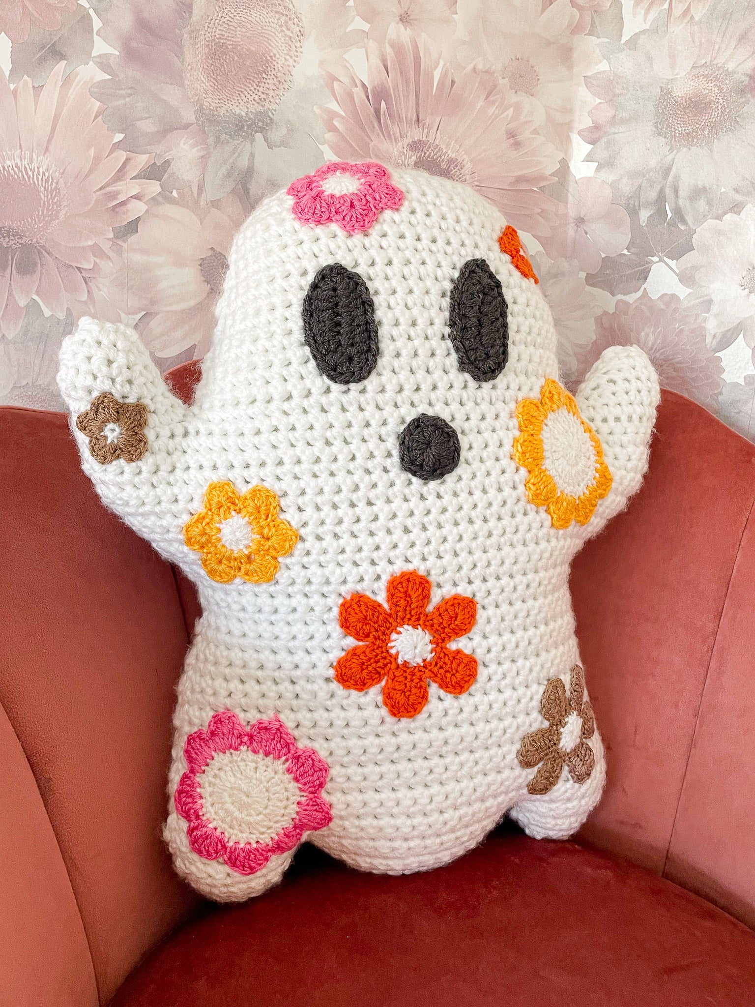 Side view of a ghost shaped pillow with flowers all over it sitting on a pink velvet chair with a floral wall paper in the back.