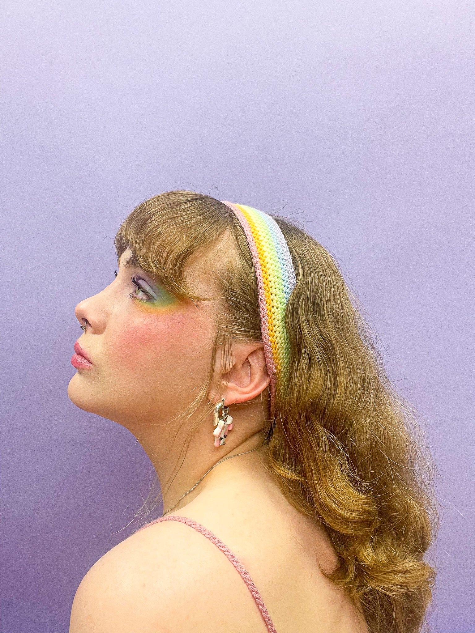 Side view of a woman posing wearing a pastel rainbow headband In her hair with matching eyeshadow sitting in front of a purple background.