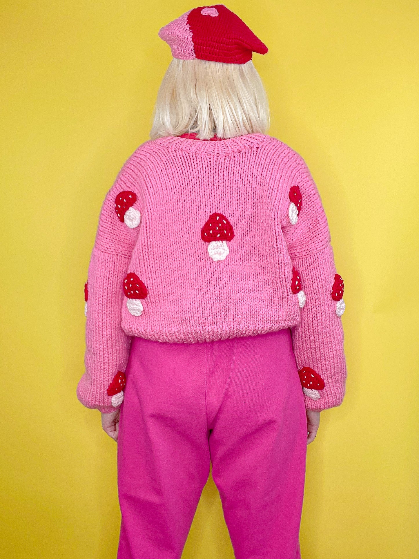 Back view of a woman wearing a pink knit cardigan with red mushrooms all over with a pink and red beret and pink overalls standing in front of a yellow background.