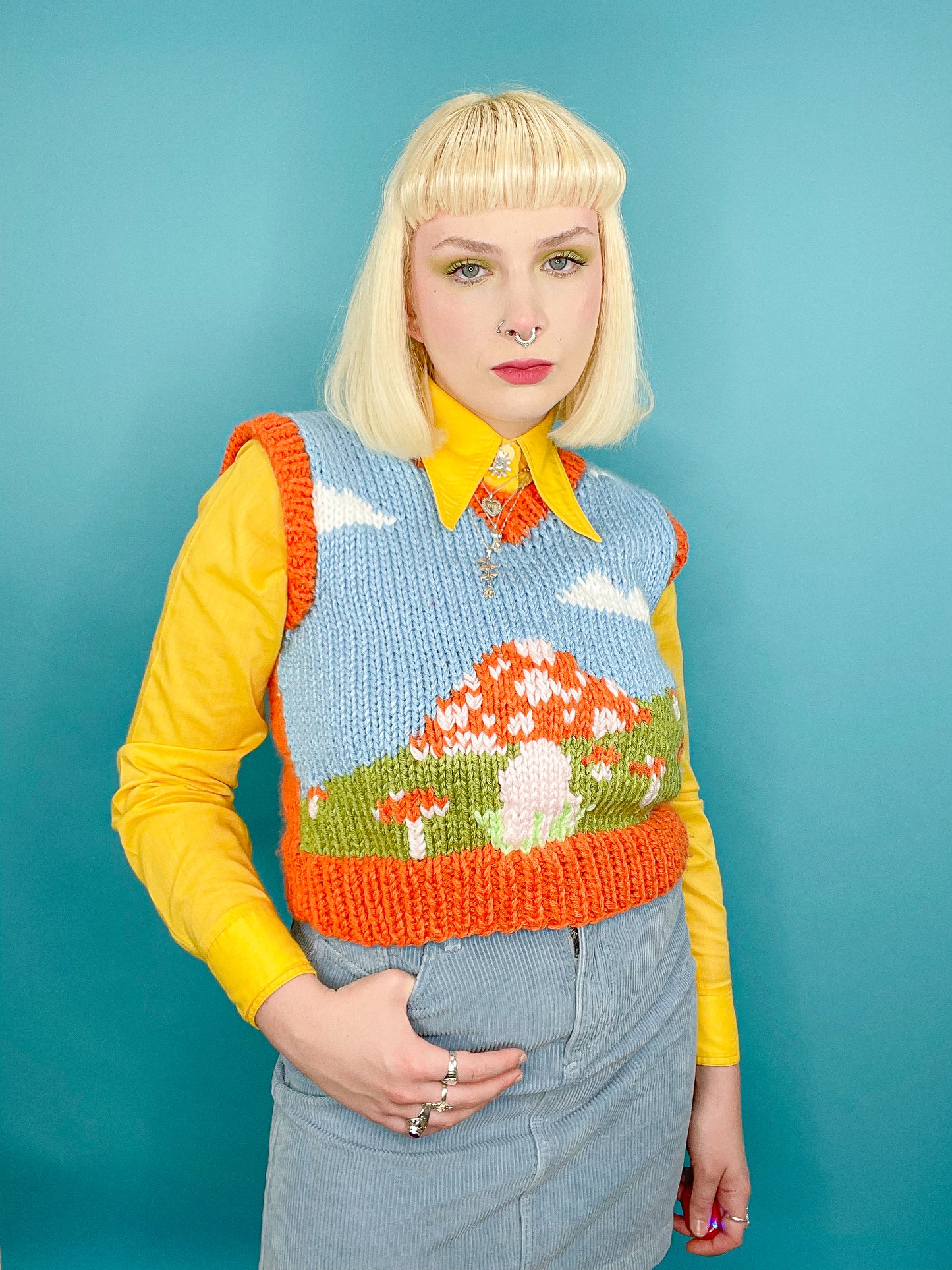 A woman posing wearing a hand knit vest with a mushroom fields design with a baby blue background and orange mushrooms and edges.