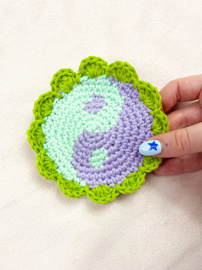 A hand holding a coaster resembling a yin yang in mint green and lilac with a bright green frilly edge.