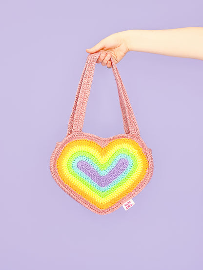 A hand holding a pastel rainbow shoulder bag shaped like a heart in front of a lilac background.