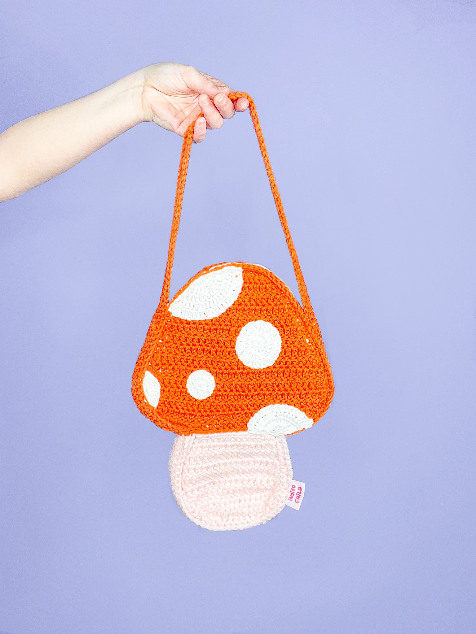 A hand holding an orange crochet bag shaped like a mushroom with appliqué dots and an Indigo Child tag sewn into the side.