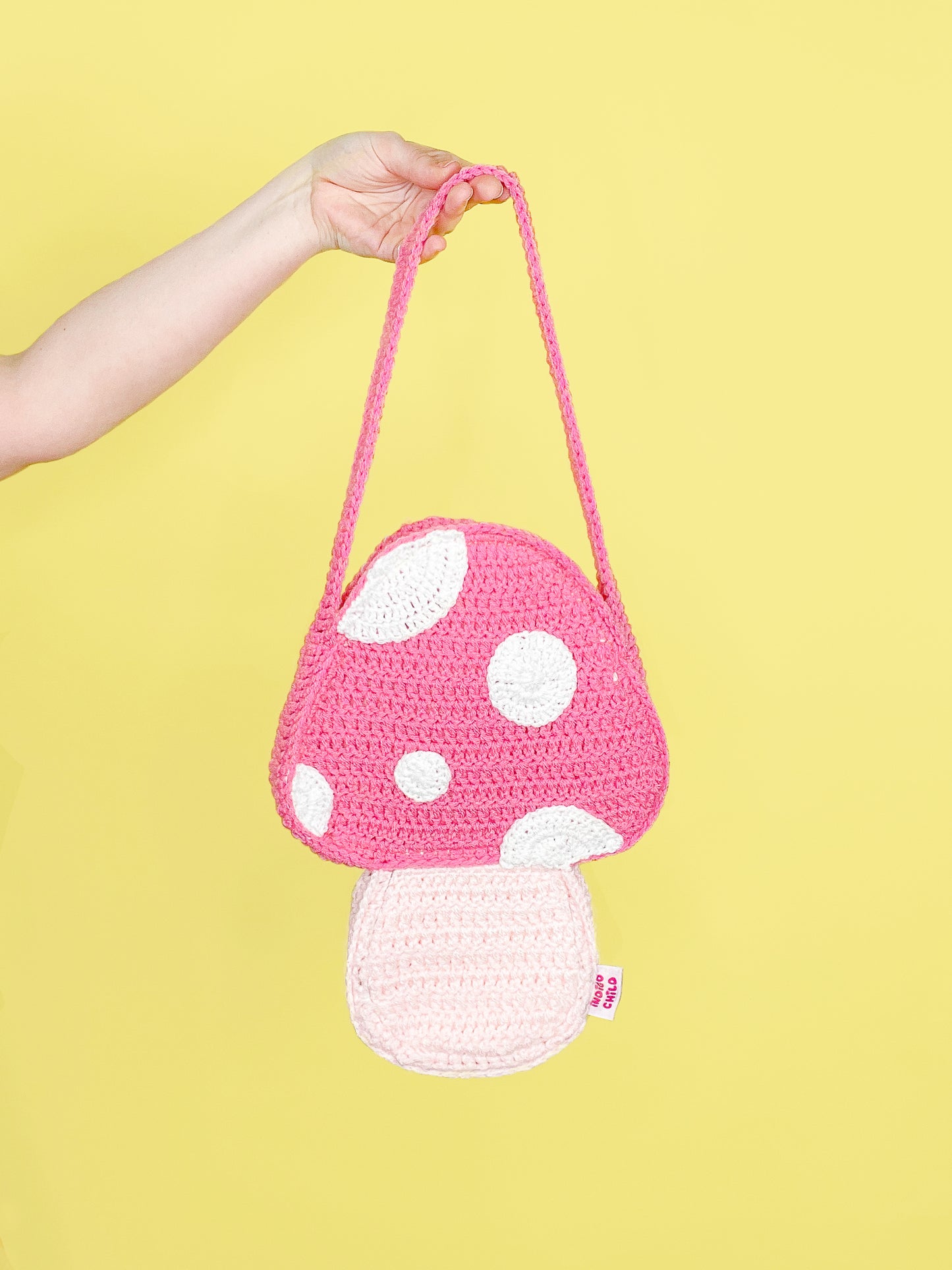 A hand holding a crochet bag shaped like a mushroom in pink in front of a yellow background.